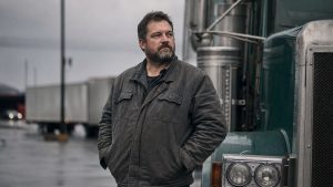 CDL disqualification — a bearded truck driver in a gray work jacket standing by a green long-nose semi truck on a rainy truck stop lot