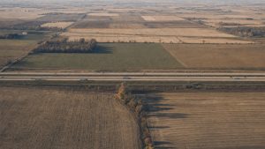 truck driver salary by state — aerial view of an interstate crossing harvested Midwest farmland with semi trucks on the road in late afternoon light